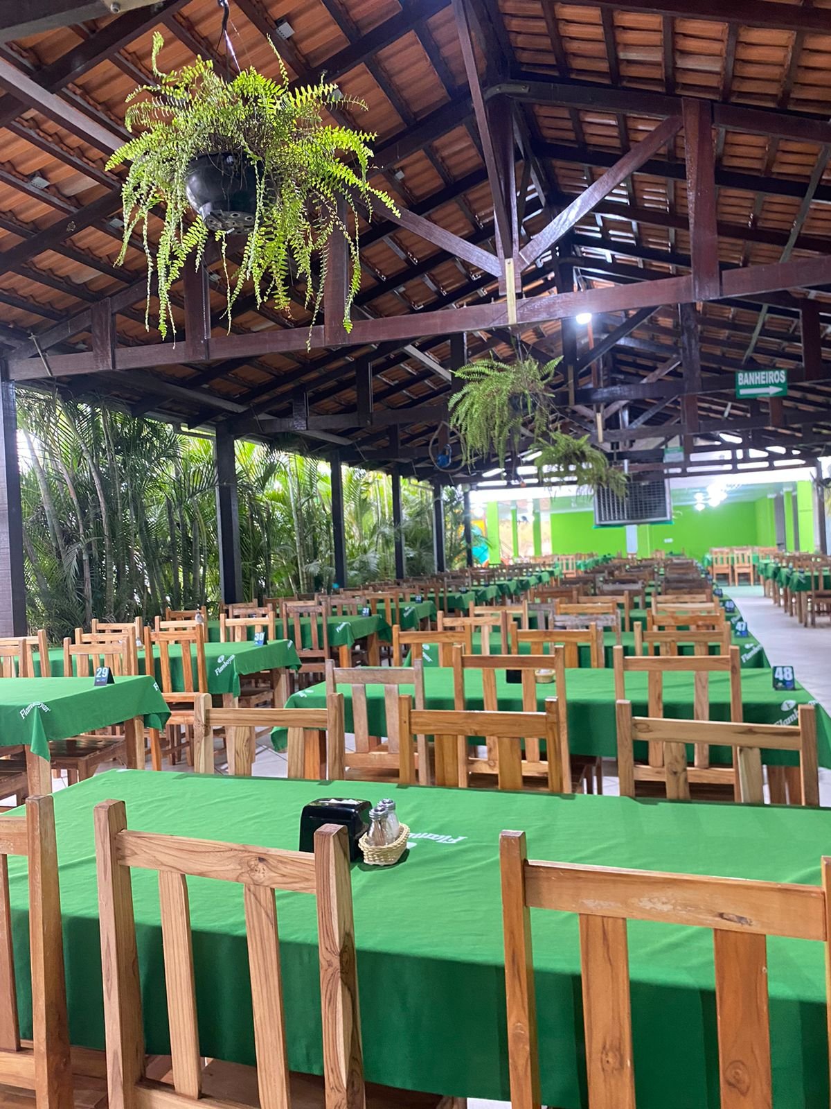 Salão rústico com telhado de madeira, mesas de toalha verde e samambaias