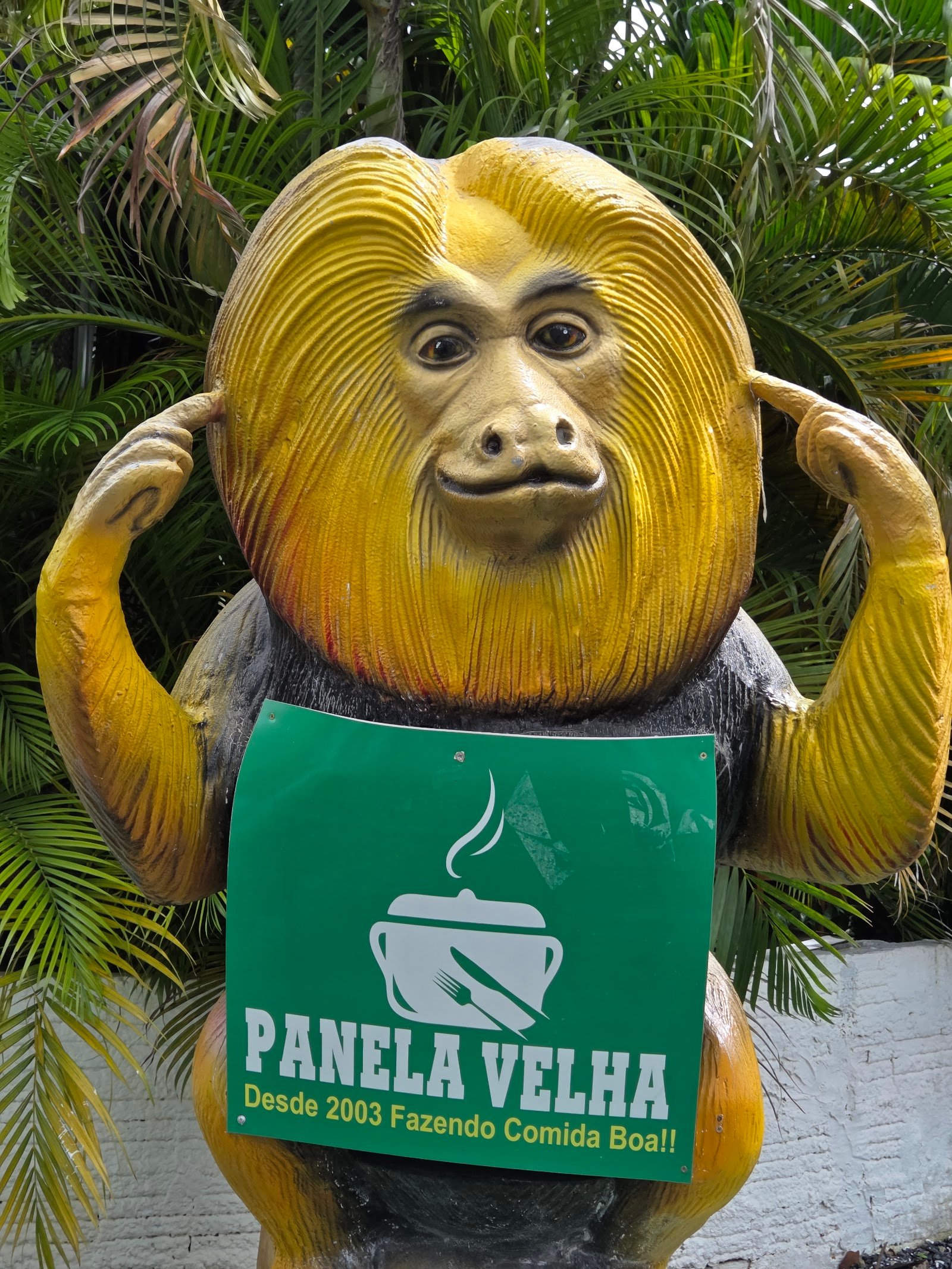 Estátua do mascote mico-leão-dourado segurando a placa Panela Velha, desde 2003 fazendo comida boa